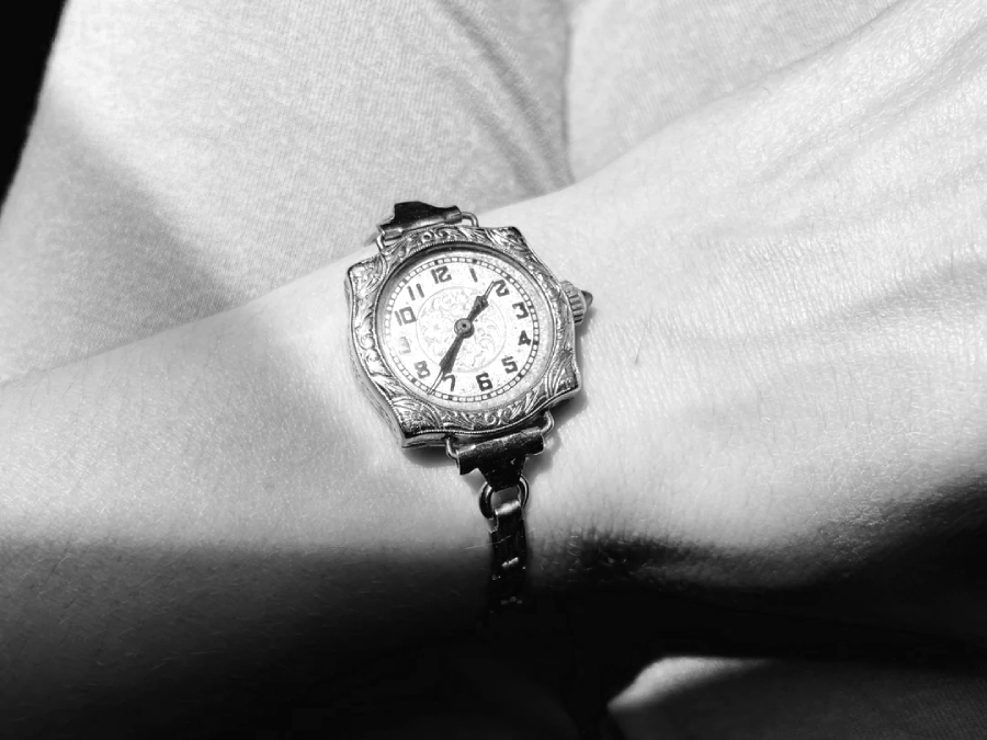 A close-up black and white photo of a wrist wearing a vintage, ornate analog watch with a round face and a decorative band. The hand and wrist are resting on fabric, possibly clothing.