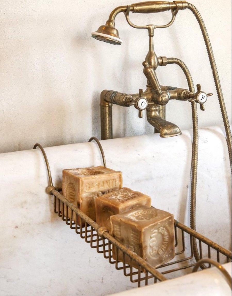 Vintage brass bath faucet with handheld shower and two white porcelain handles over a marble tub. Three large blocks of brown soap rest on a wire rack above the tub.