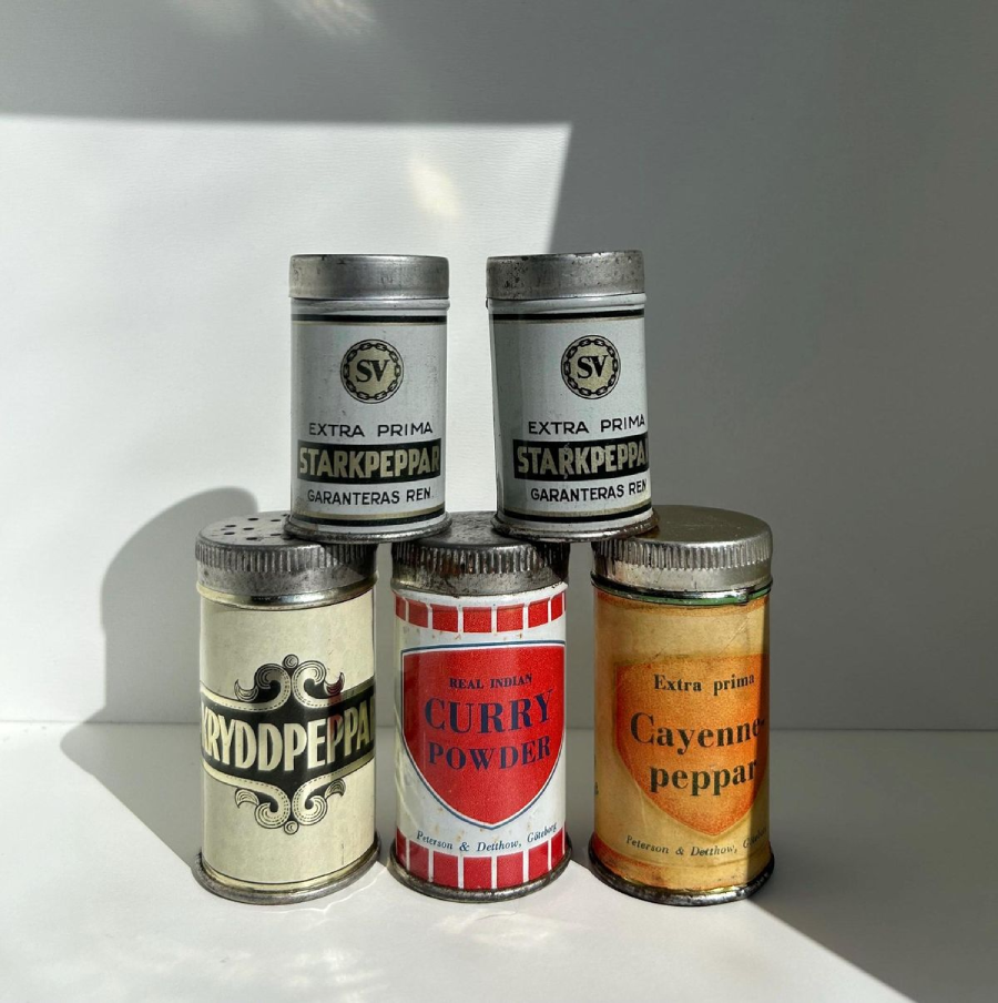 Five vintage spice tins with worn labels are arranged in sunlight against a white background. The labels read "Starkpeppar," "Ryddpeppar," "Curry Powder," and "Cayenne Peppar.