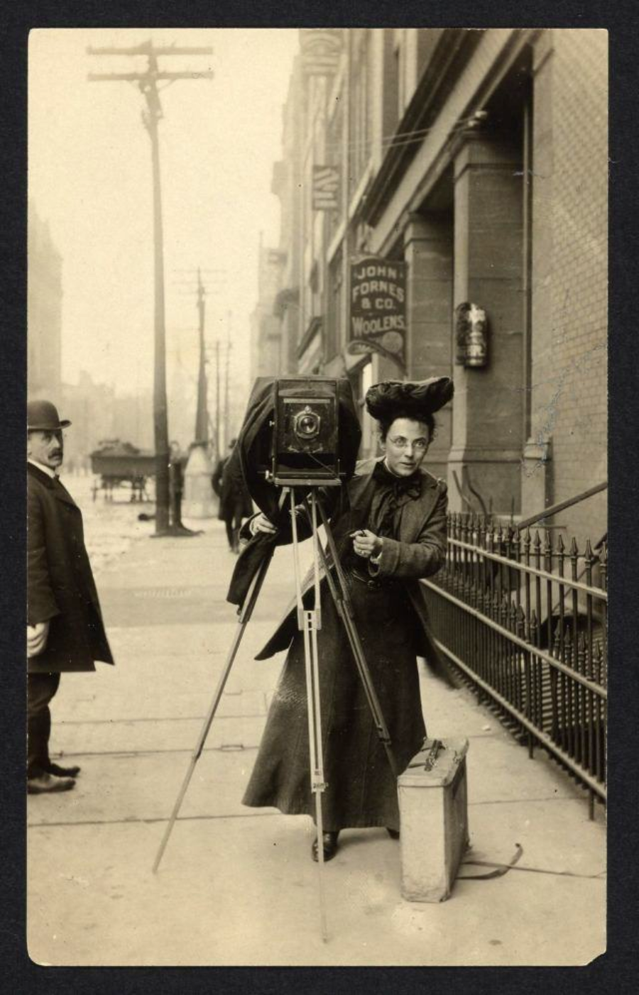A woman in early 20th-century clothing operates a large box camera on a tripod on a city sidewalk. A man in a bowler hat walks nearby. Buildings, a storefront sign, and a fence are visible in the background.
