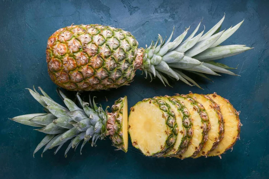 A whole pineapple is lying on a dark surface, with half of it sliced into round pieces, showing the yellow flesh and green spiky crown.