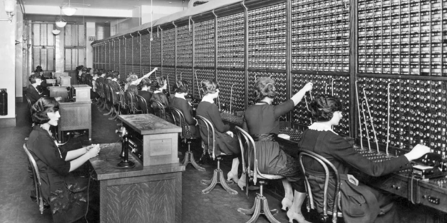 Black and white photo of women working at a large, old-fashioned telephone switchboard, plugging cables in and out to connect calls, sitting in a row and wearing similar dark dresses.