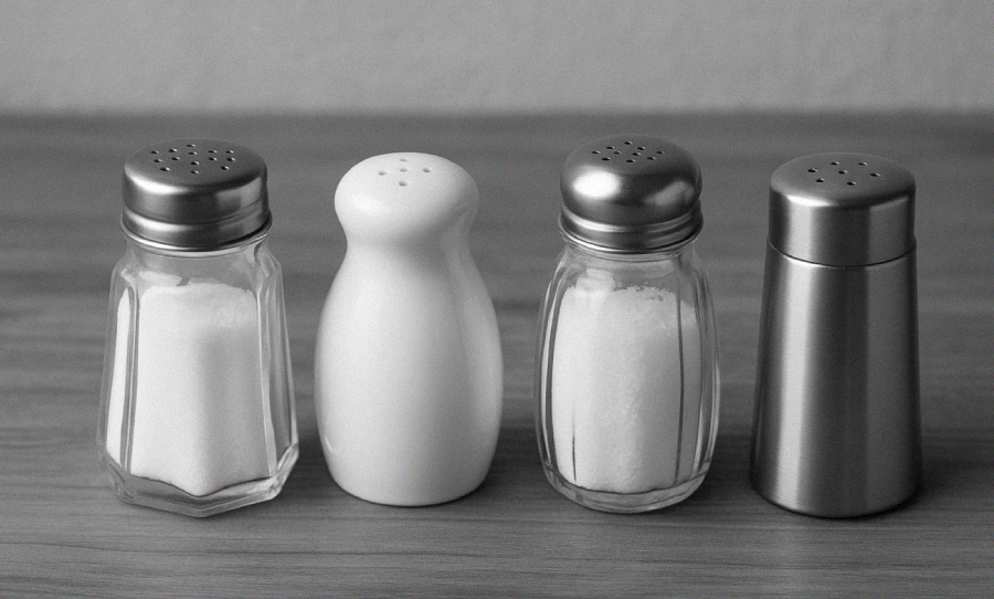 Four salt shakers of different shapes and materials are lined up on a wooden surface; two are glass with metal tops, one is ceramic, and one is metal.