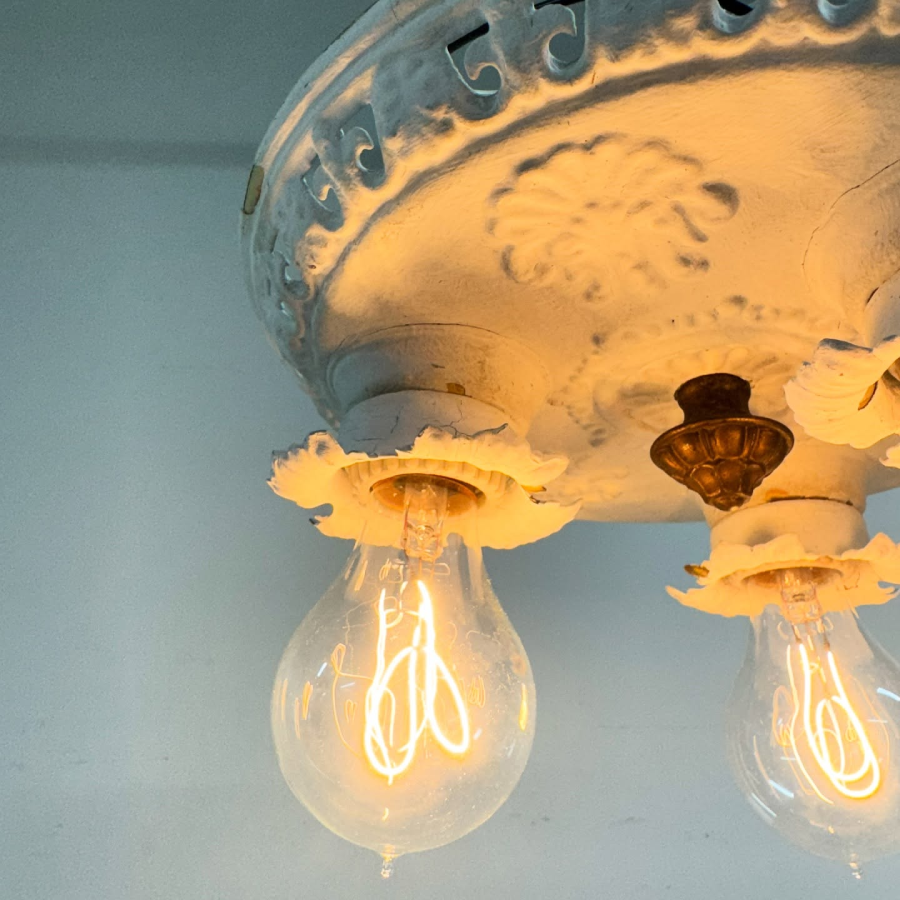 A close-up of a vintage, ornate white ceiling light fixture with exposed glowing Edison-style light bulbs, set against a pale blue background.