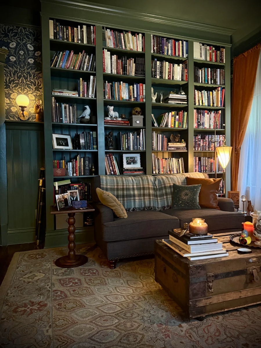 A cozy library with floor-to-ceiling bookshelves filled with books, a gray couch with plaid cushions, a wooden trunk used as a coffee table, warm lamps, candles, and a patterned rug. Natural light filters through sheer curtains.