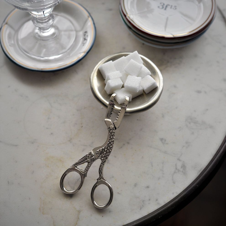 A pair of ornate sugar tongs shaped like a bird holds a small dish with sugar cubes, resting on a marble tabletop next to a glass and stacked saucers.
