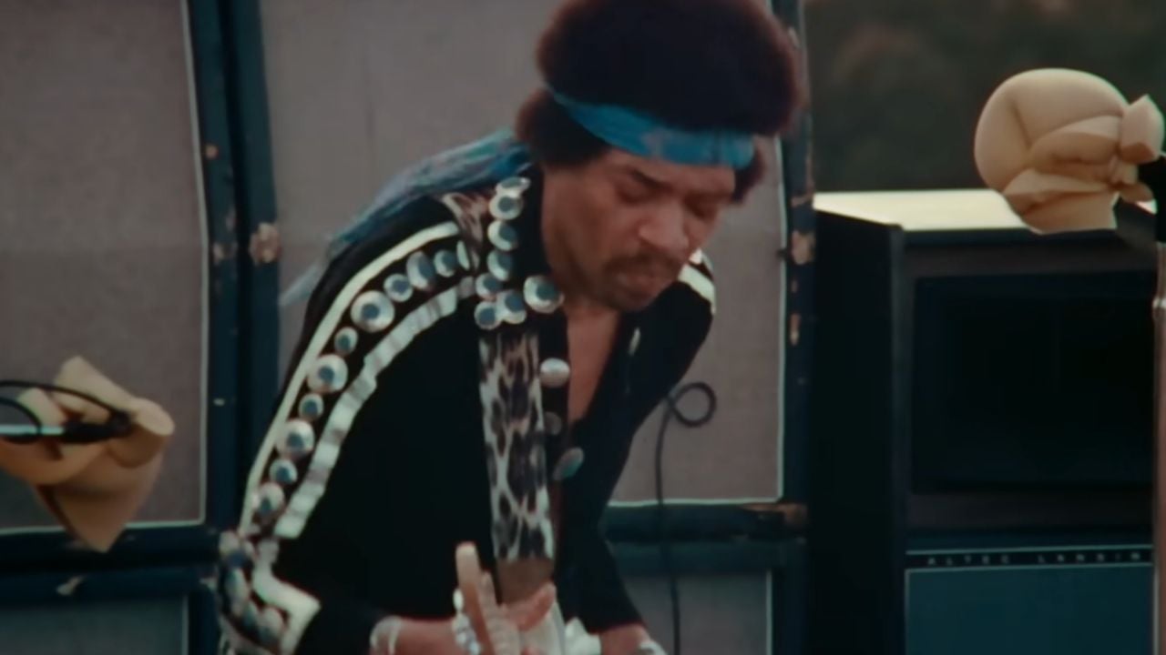 A musician wearing a blue headband and a black outfit with silver discs plays an electric guitar outdoors, with speakers and equipment visible behind him.