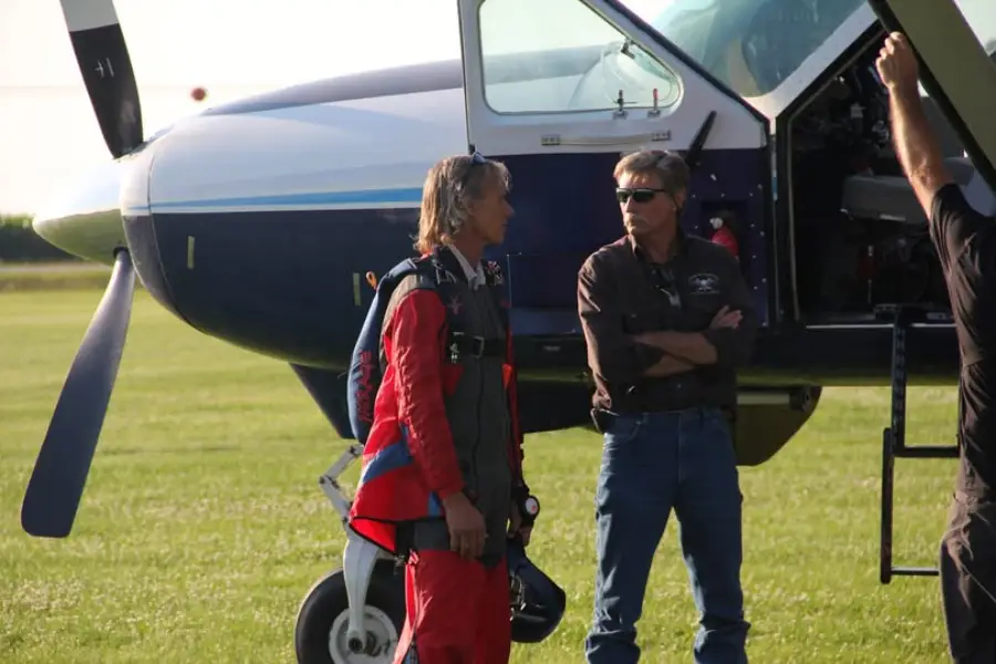 Two men stand beside a small propeller airplane on a grassy field; one wears a red jumpsuit and parachute, the other wears sunglasses and casual clothes, with the plane door open behind them.