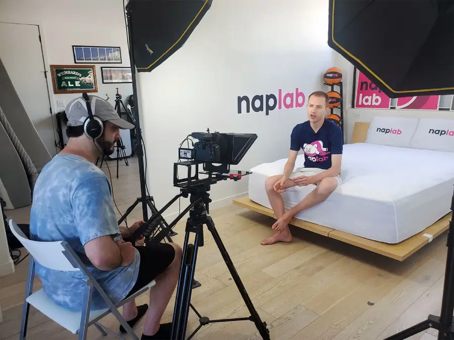 A man sits barefoot on a bed in a studio with “naplab” branding while another man, wearing headphones, films him using a video camera and professional lighting.
