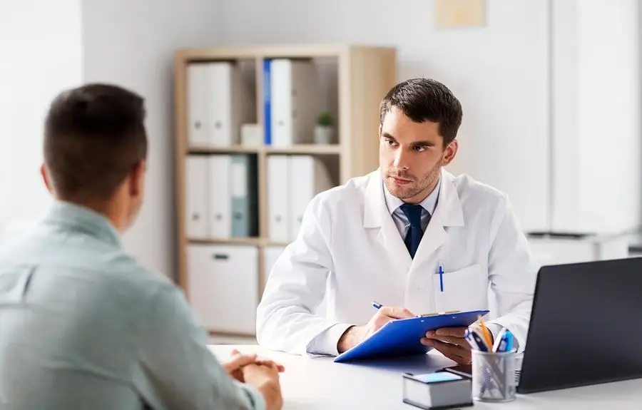 A doctor in a white coat sits at a desk, holding a clipboard and pen, attentively listening to a patient across from him in an office setting.