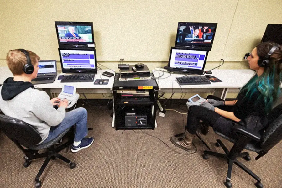 Two people sit at desks with headphones, typing on devices while watching news videos on dual computer monitors, possibly transcribing or captioning the content. Various equipment and cables are on the table between them.