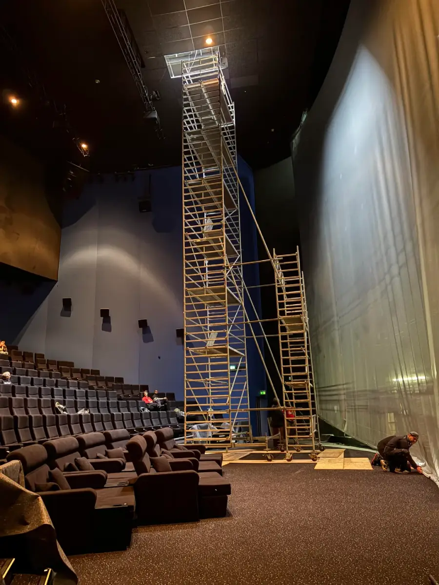 A tall scaffold tower stands on a theater stage beside a large curved screen, with empty seats in the audience. Two people are working near the stage floor, and another person sits in the theater seats.