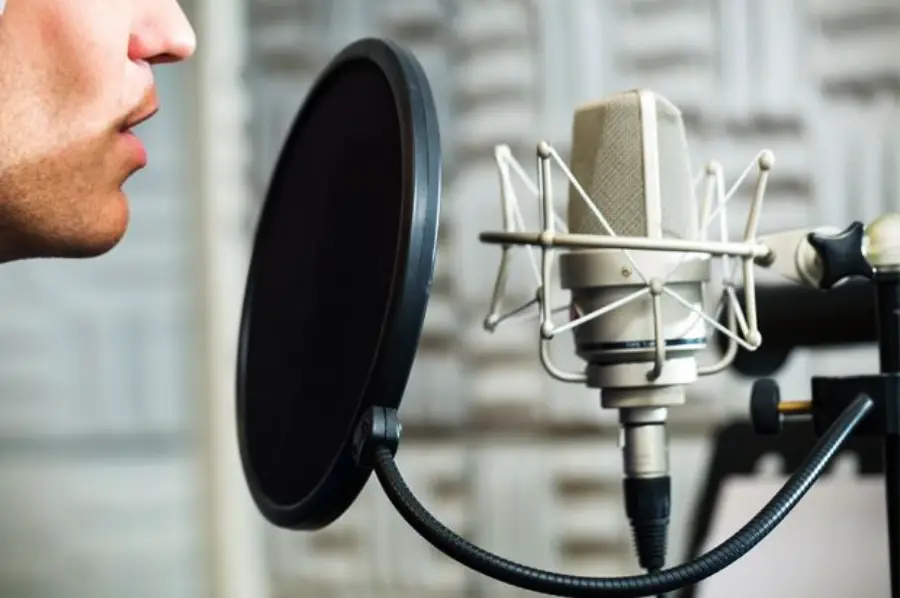 A person speaking into a professional microphone with a pop filter in a recording studio, with acoustic foam panels in the background.
