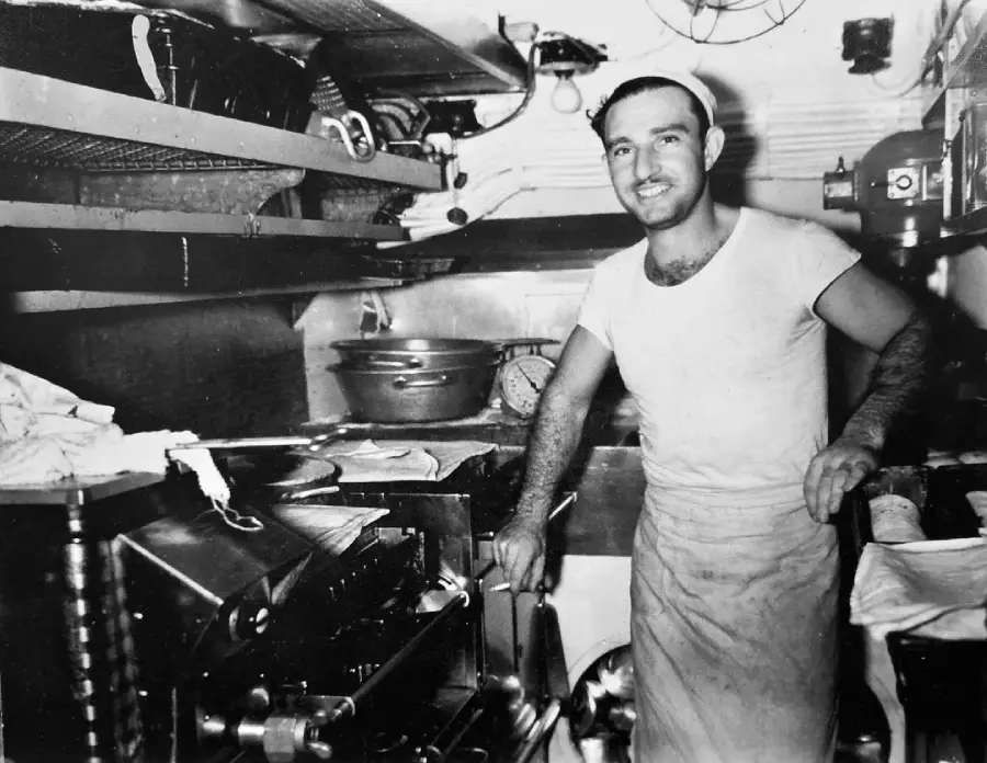 A smiling man wearing a white T-shirt, apron, and cap stands in a small, busy kitchen surrounded by shelves, kitchen utensils, and cooking equipment.