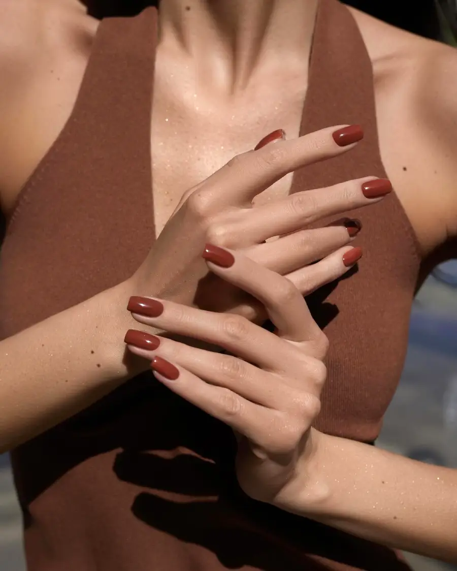 A person wearing a sleeveless brown top poses with hands gracefully crossed, showing off long, almond-shaped nails painted in a glossy brown color.