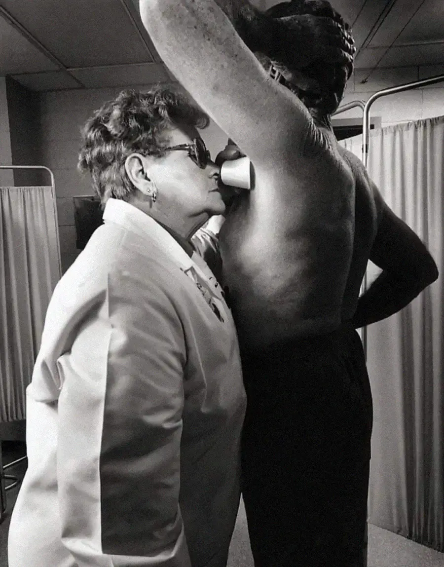 A woman in a white coat smells the armpit of a shirtless person raising their arm in a clinical setting, possibly conducting an odor test; privacy screens are visible in the background.