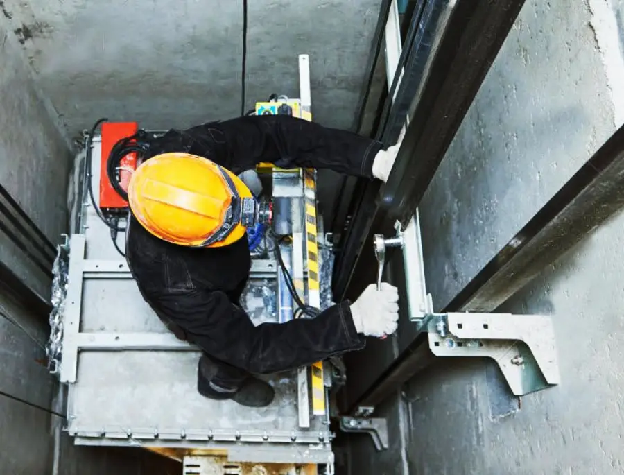 Worker wearing a yellow hard hat and black clothing repairs or maintains an elevator mechanism inside an elevator shaft, using tools on metal components. The scene is viewed from above.