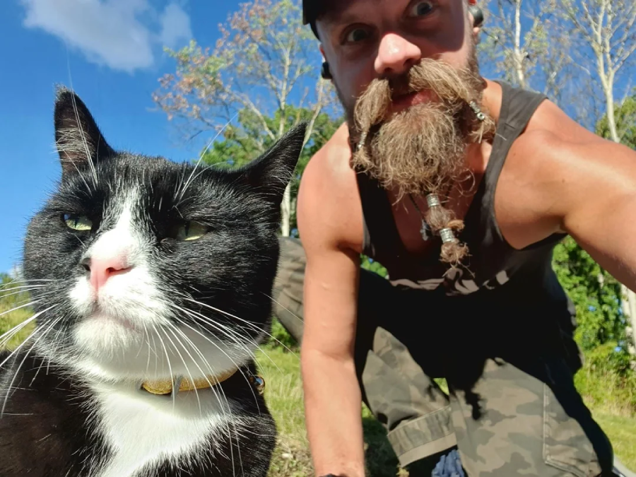 A man with a bearded, braided mustache and a black tank top crouches outdoors next to a black and white cat wearing a yellow collar under a blue sky.