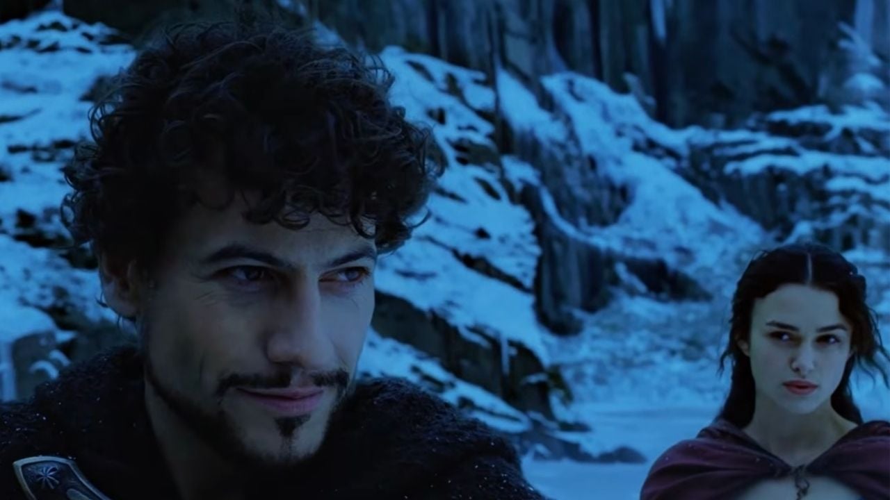 A man with curly hair and a trimmed beard stands in snowy mountains, looking ahead, while a woman with dark hair and a serious expression stands behind him. The scene is cold and dramatic.