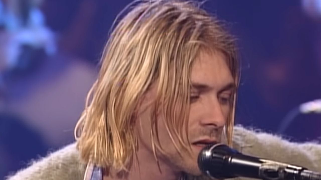 A person with shoulder-length blonde hair sings into a microphone on stage, wearing a light-colored sweater. The background is dimly lit with soft colors.
