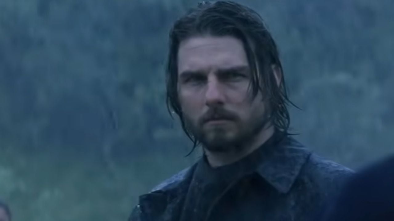 A man with long, wet hair and a beard stands outside in heavy rain, wearing a dark coat and looking serious.