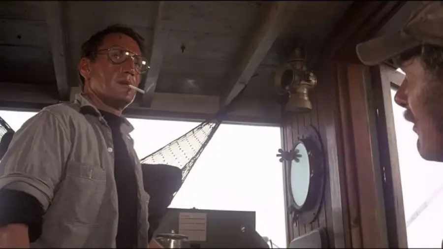A man with glasses and a cigarette in his mouth stands inside a boat's cabin, looking toward another man in a cap. Nautical equipment and a round window are visible in the background.
