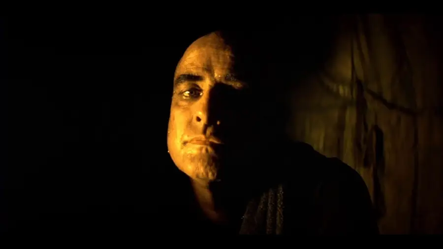 A man with a shaved head sits in dim, dramatic lighting, half his face illuminated and the other half cast in deep shadow, creating a moody and intense atmosphere.