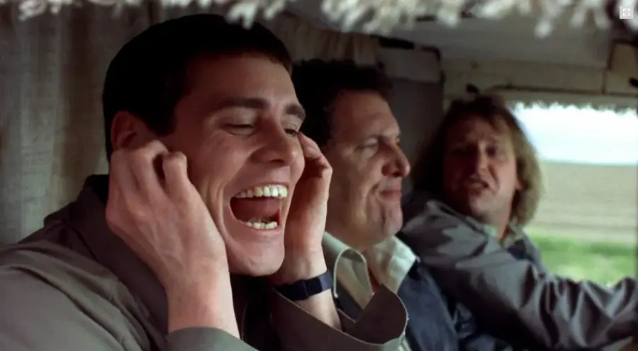 Three men sit in the front seats of a vehicle. The man on the left smiles widely with his mouth open and covers his ears, while the other two look forward. They appear to be in the middle of a conversation or joke.