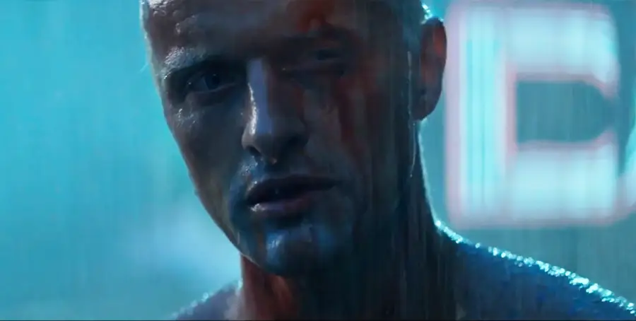 A close-up of a man standing in the rain with water streaming down his face, illuminated by blue neon light, looking intensely ahead with a somber expression.