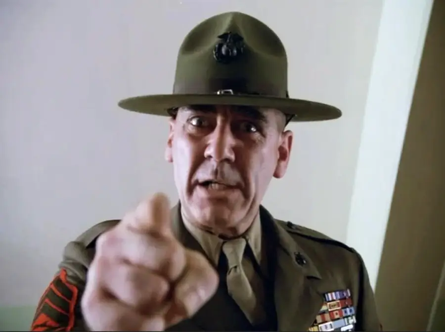 A stern military drill instructor in uniform, wearing a campaign hat, points directly at the camera. Medals and badges are visible on his chest, and he appears to be shouting or giving a command.