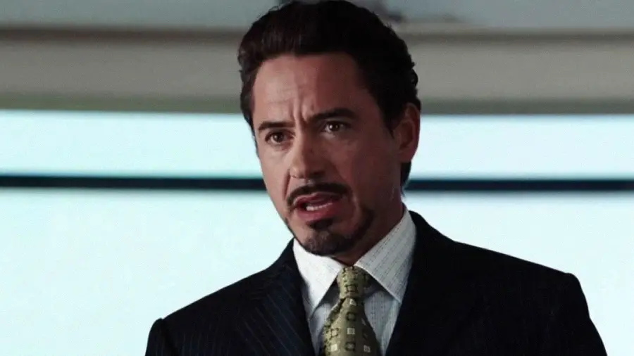 A man with short dark hair and a goatee, wearing a dark pinstripe suit, light shirt, and patterned tie, stands indoors and speaks with a serious expression.