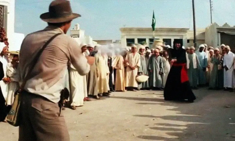 A man in a fedora and brown clothes aims a gun at a swordsman dressed in black and red, while a crowd of people in traditional Middle Eastern attire watches in a sunny outdoor market setting.