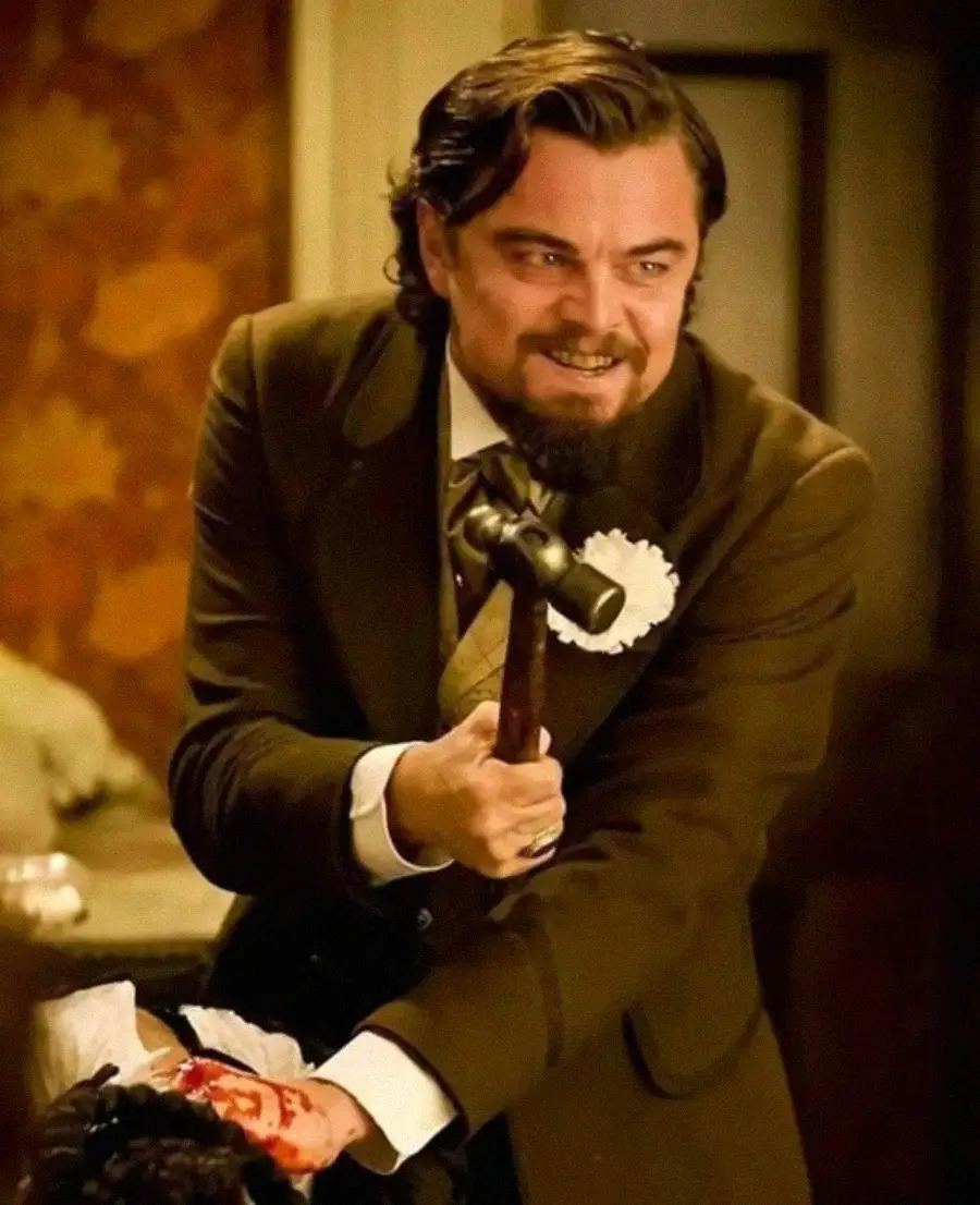 A man in a brown suit with a flower on his lapel holds a hammer menacingly, grinning with intensity. His hair is slicked back and he stands over someone whose hand appears injured and bloody.