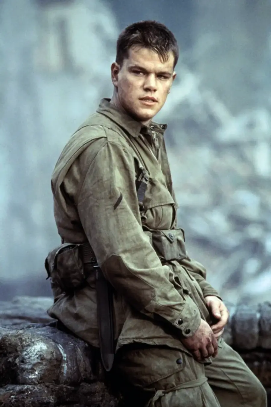 A young man in a worn, olive-green World War II military uniform sits on a rock, looking into the distance with a serious expression. The background is smoky and rubble-filled, suggesting a war zone.