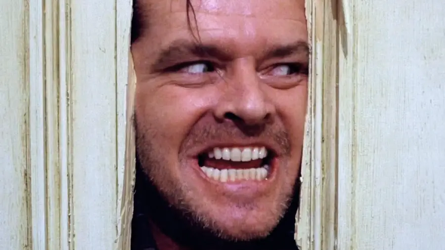 A man with a wild expression smiles menacingly through a broken wooden door, his face framed by jagged edges, eyes wide and teeth bared.