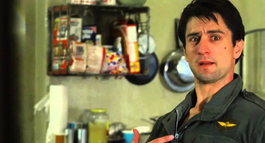 A man with short dark hair and a serious expression stands in a kitchen, pointing at himself while wearing a green jacket. Shelves with food items and kitchen utensils are visible in the background.