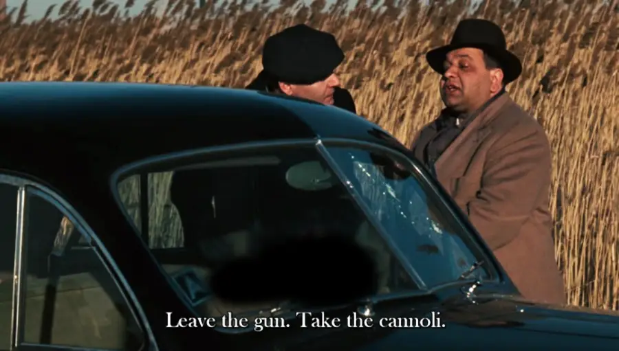 Two men stand beside a black car in a field of tall grass, one speaking to the other. Subtitles read: "Leave the gun. Take the cannoli.