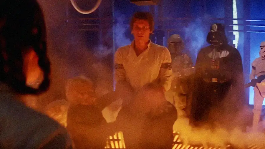 A man in white clothing is restrained with his hands bound, surrounded by armored guards and a black-clad figure with a helmet, in a smoky, brightly-lit industrial setting.