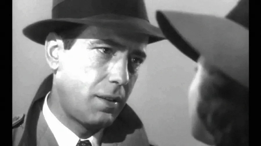 A man in a fedora and trench coat looks intently at a woman wearing a hat, both in black and white, creating a classic film noir atmosphere.