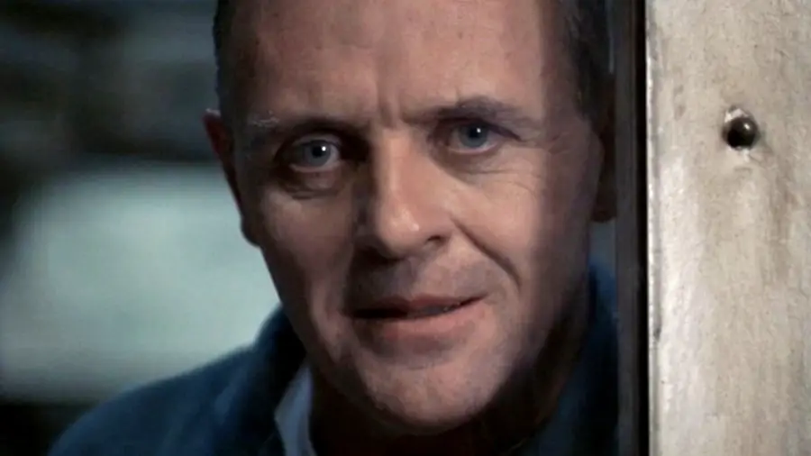 A middle-aged man with light skin, blue eyes, and a slight smile looks through a glass window, partially framed by a wooden panel, in a dimly lit setting.