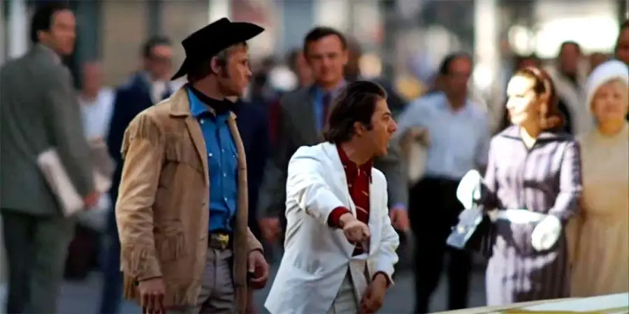 Two men stand on a busy city street, one in a cowboy hat and tan jacket, the other in a white jacket gesturing and shouting, while blurred pedestrians walk by in the background.