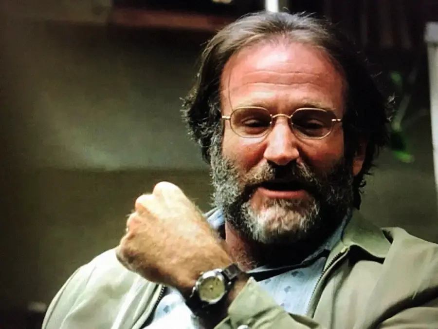 A man with glasses and a beard, wearing a light jacket and watch, gestures with his right hand while talking. The background is out of focus.