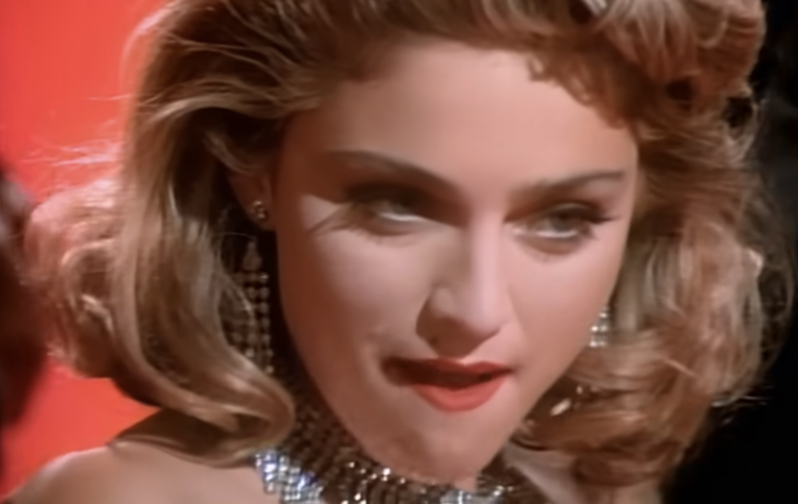 A woman with wavy blonde hair, red lipstick, and glamorous earrings looks forward with a sultry expression against a red background.