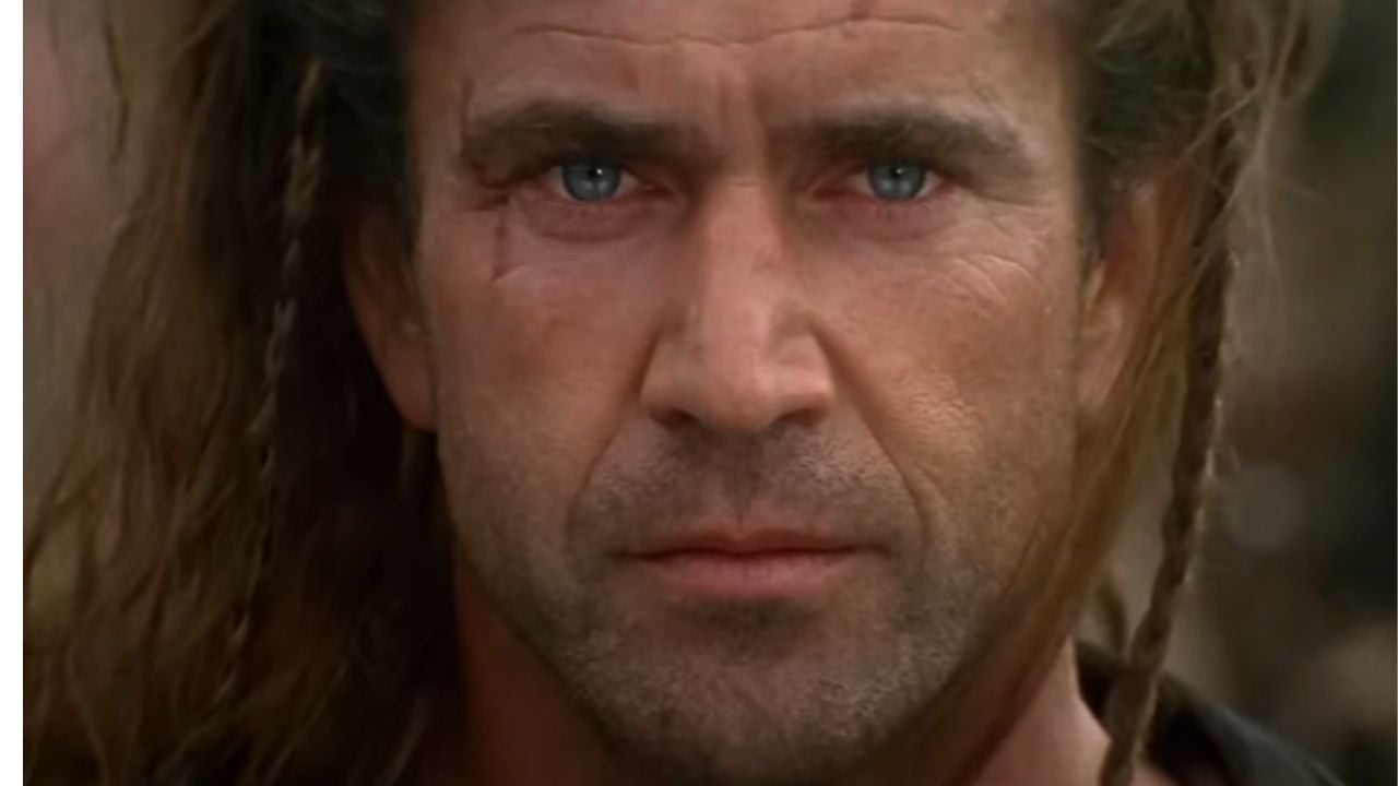 A close-up of a man with long, tousled brown hair, blue eyes, and a serious expression. He has a scar on his cheek and is outdoors in natural light.
