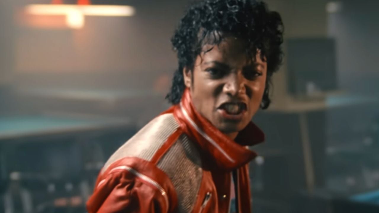 A person with curly hair wearing a red jacket looks intensely at the camera in a dimly lit room.