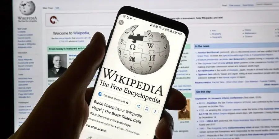 A hand holding a smartphone displaying the Wikipedia homepage, with a computer screen in the background also showing the Wikipedia website.