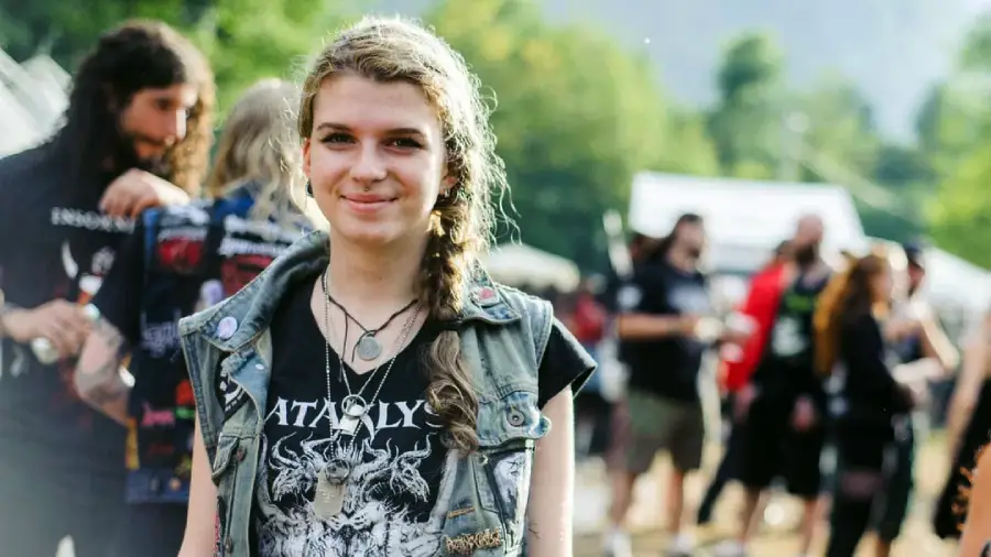 A young woman with wavy hair and a braid smiles at an outdoor event. She wears a metal band T-shirt, a vest with patches, and layered necklaces. Blurred people and greenery are visible in the background.