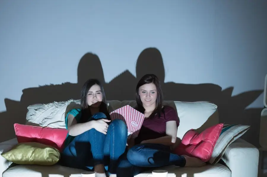 Two young women sitting on a couch with colorful pillows, watching TV in a dimly lit room, sharing a striped popcorn container, their shadows cast on the wall behind them.