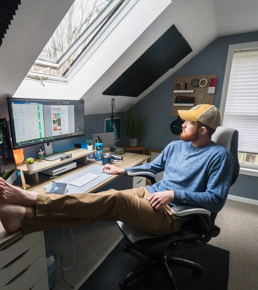 A man wearing a cap and blue sweater sits at a desk in a home office with feet up, looking out a skylight window. Multiple monitors, a keyboard, and office supplies are on the desk; daylight streams in.