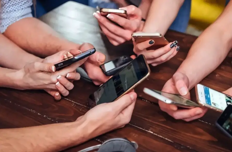 Several people sit around a wooden table, each holding and using a smartphone, with only their hands and phones visible.